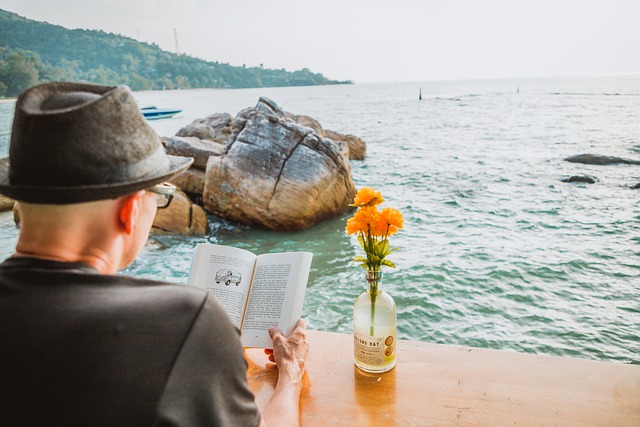 Homem de chapéu e óculos a ver um livro em frente ao mar e a pensar quanto ganha um escritor em Portugal.