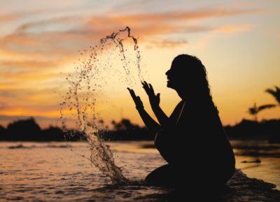 Sombra de mulher no mar, a atirar água ao ar, sorridente e com o pôr-do-sol ao fundo a pensar em como desligar do trabalho nas férias.