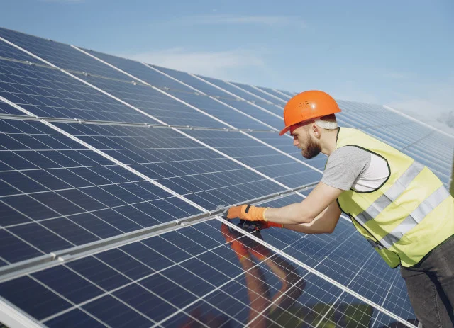 Instalador de energia solar e eólica a trabalhar em painéis solares com capacete laranja e colete amarelo.